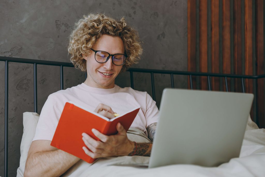 Person mit Brille sitzt mit rotem Notizbuch und Laptop im Bett.